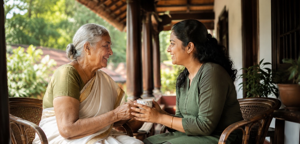 A caregiver in Kerala sitting beside an elderly woman on a veranda, handing her a cup of tea while the two look at each other and smile